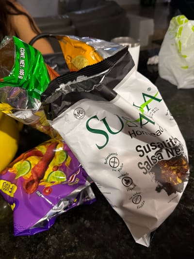 The image shows a pile of opened snack bags on a dark countertop. Visible brands and types of snacks include "Susalitas Salsa Negra", "Takis Fuego" (specifically, "EXTREMO" flavor), and another green bag labeled "CONT. NET. 185 g" of an unidentifiable snack. The scene appears to be indoors. The OCR suggests the presence of the "MK PAREVE" symbol, and information suggesting the product is from Mexico, specifically referring to ingredients and packaging. The image does not contain enough information to determine its exact location in Santa Clara Ocoyucan, Mexico.
