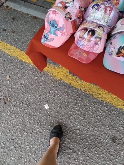 This is a high-angle, full shot of several baseball caps with cartoon characters and K-Pop idols displayed on a red cloth on the ground next to an asphalt surface with a yellow line. The caps feature popular characters like Minnie Mouse and Stitch, as well as images of K-Pop groups. Some of the text visible on the caps includes "HUNTRIX" and "SKIKBOYS." A person's leg and foot, wearing a black espadrille, are visible in the lower right corner, suggesting the caps are for sale. The setting appears to be outdoors, possibly a market or street stall, with the asphalt and yellow line indicating a roadside or pedestrian area. The lighting suggests it is daytime, and the weather appears to be clear.
