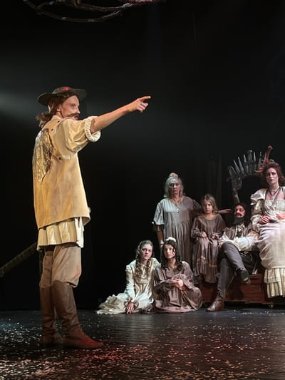 This image depicts a stage performance occurring at a theater or cultural center in Novoshakhtinsk, Russia. Seven individuals in period-style costumes are illuminated by spotlights on a dark stage, which features scattered light-colored fragments on its surface. On the far left, a male actor with a mustache and long, dark hair, dressed in a light-colored shirt, a tan vest with a metallic design, tan trousers, and dark brown lace-up boots, stands in profile. He extends his right arm directly forward, pointing with his index finger. To his right, a group of six other individuals is arranged. An older woman with light hair and a pale face, wearing a long grey dress and necklace, stands centrally. Beside her, a young girl in a dark brown dress stands with her gaze directed forward-left. In the front row, two young women are seated on the floor, both wearing long dresses (one cream, one dark brown) and looking forward-left. On the right, a man with a full dark beard and a woman with elaborate reddish headwear are seated on a dark wooden chest-like prop. Both are dressed in light-colored period attire and display serious expressions. Overhead, an abstract, dark, skeletal-like structure is partially visible, and a similar jagged prop serves as a backdrop behind the seated group on the right.