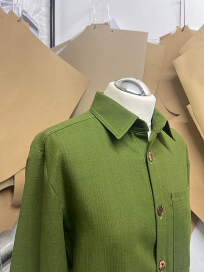A green button-up blazer hangs on a mannequin, surrounded by tan and white paper, likely pattern pieces, against a white wall and possibly a tiled floor. The environment suggests a design or sewing studio. There is no discernible information about location in the city of Kirklees, United Kingdom, or people present in the image. The focus is solely on the blazer and the surrounding design-related objects.