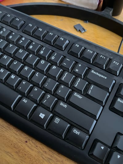 The image shows a close-up, high-angle shot of a black computer keyboard resting on a wooden surface. The keyboard is angled, with the top edge towards the viewer's upper right. Several keys are clearly visible, including function keys F2 through F12, number keys, letter keys, and modifier keys such as Alt, Shift, Ctrl, and the Windows key. Some keys have symbols printed on them, such as percentage, ampersand, asterisk, and punctuation marks. A small, gray plastic piece, possibly a keycap or a part of a computer accessory, is visible on the wooden surface to the left of the keyboard. The background is out of focus, showing a dark, curved object, likely part of a monitor stand or a similar piece of computer equipment. The lighting suggests an indoor environment. There are no people or animals in the image. The scene is static and appears to be a detail shot of computer hardware. There is no information in the image to suggest the location is the city of Kedawung, Indonesia.
