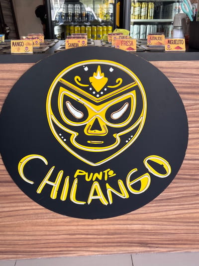 A circular sign displays a stylized wrestler mask logo in yellow and white outlines on a black background. The name "CHILANGO," and the word "PUNTO" are written in yellow, with a neon-like effect. The sign is mounted on a wooden surface. To the left, a refrigerator shelf holds various canned and bottled goods with labeled price tags displaying flavors such as "MIGUELITO," "AJONJOLI," "FRESA," "SANDIA," and "MANGO."