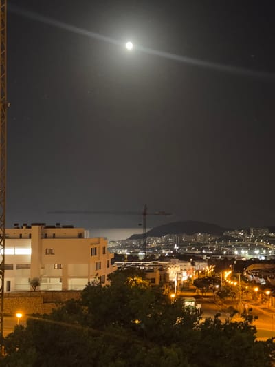 The image is a night-time photograph taken from an elevated position, likely a balcony or window, overlooking a city. A bright, full moon is visible in the dark sky, with a distinct light beam emanating from it diagonally across the upper portion of the frame. The lower half of the image shows a city with numerous lights, suggesting a populated urban area.

In the foreground, a tall yellow structure, possibly part of a construction crane or scaffolding, is visible on the left side. Below this, a modern-looking building with white facades and dark windows is prominent. A small olive tree stands in front of this building. Further into the scene, a road with streetlights illuminates a green vehicle, possibly a bus, and several other cars. More buildings and lights extend into the mid-ground and background, with a hilly landscape and a distant city skyline visible. Another construction crane is present in the mid-ground. The overall impression is of an urban landscape at night under a bright moon.

The location is identified as Finestrat, Spain.