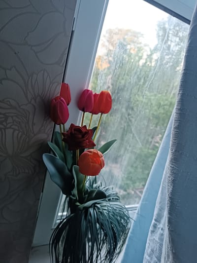 An artificial floral arrangement featuring several tulips and a rose is placed in a vase on a windowsill. The flowers are red, pink, and orange. The vase is dark green and appears to have a feathery or spiky texture at the top. Behind the flowers, a window looks out onto a blurred background of trees and foliage, suggesting an outdoor environment. A light blue curtain is visible to the right of the window. The wallpaper on the left side of the image has a subtle floral pattern. The scene appears to be indoors, likely in a residential setting. The location is not specified as being in Chirchik, Uzbekistan.