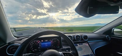 A forensic examination of the image reveals a driver's perspective from inside a modern Mercedes-Benz vehicle, traveling on a multi-lane highway near Zavoy, Bulgaria.

The interior foreground shows a black leather-wrapped steering wheel, with a digital instrument cluster behind it. The speedometer indicates a current speed of 132 km/h, with a "LIM 110" warning, suggesting the vehicle is exceeding a detected speed limit of 110 km/h. The odometer reads 109868 km, with a trip meter showing 5688.1 km. The time is 19:05, and the ambient temperature is 25.0 °C. The transmission is in "D 9" (Drive, 9th gear), and the low beam headlights are active. To the right, a large, vertically oriented infotainment touchscreen displays a navigation map with local topography, including a body of water. Bulgarian text is visible on this screen, alongside "Телефон S23 Ultra," indicating a connected Samsung S23 Ultra phone. The dashboard features three circular air vents with metallic bezels and a strip of blue ambient lighting. A rearview mirror is mounted centrally at the top of the windshield. An obscure inscription, possibly "FWTA," is visible on the upper left of the windshield.

The exterior view, seen through the windshield, depicts a rural landscape under a partially cloudy sky. The sun, positioned towards the left, pierces through the clouds, casting visible crepuscular rays. The highway stretches into the distance, with a single vehicle visible far ahead. Green fields border both sides of the road, separated by a guardrail. In the background, rolling mountain ranges extend across the horizon, with a distinct, lighter peak visible on the far right. Red and white striped marker poles are visible along the right side of the highway. The overall scene suggests late afternoon or early evening conditions. No individuals are visible within the frame.
