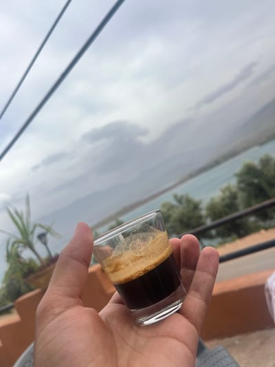 The image, captured from a tilted perspective, primarily features a person's right hand holding a small, clear glass. The glass contains a dark coffee beverage, likely an espresso shot, topped with a distinct, thick layer of light brown crema.
The scene is set outdoors, offering a panoramic view in the background. An expansive body of water, possibly a reservoir or lake, stretches across the midground towards the horizon. On the distant bank of the water, green trees are visible, indicating lush natural surroundings. Above the landscape, the sky is bright but overcast, with soft, diffused light suggesting a cloudy daytime. Two dark lines, resembling power cables or wires, diagonally traverse the upper-left portion of the frame against the sky. To the right of the hand, elements of outdoor architecture, including a terracotta-colored wall and part of a railing or fence, are visible. The setting suggests a viewpoint from a balcony or terrace, likely in Bin El Ouidane, Morocco. No text is visible in the image.