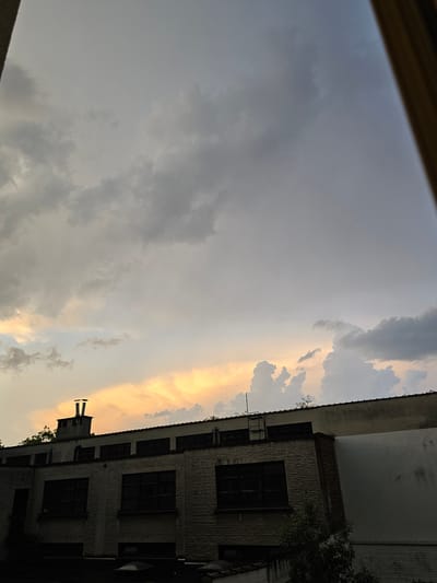 The image captures a view from a window, with a portion of a building visible to the right and the sky dominating the scene. The building is constructed of brick, has multiple windows with dark frames, and a chimney protruding from its roof. Patches of greenery, possibly trees, are visible near the building. The sky is filled with clouds, displaying a gradient from grey to orange and yellow, suggesting either sunrise or sunset. The photo appears to be taken from indoors, with a portion of the window frame visible at the top. Anderlecht, Belgium is not discernible from the visual elements.