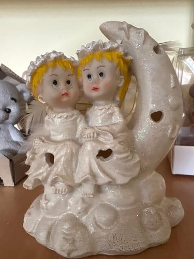 A close-up, eye-level shot focuses on a decorative figurine of two angelic children with yellow hair and white halos, seated on a crescent moon. The figurine is primarily white with glitter accents, and the moon has two heart-shaped cutouts. The children's faces are painted with blue eyes, rosy cheeks, and red lips. Feathery white accents suggest wings. The figurine rests on a cloudy base. To the left of the figurine, partially out of focus, is a small, light gray teddy bear. The background consists of a plain, light-colored wall and hints of other objects, suggesting an indoor setting. The overall scene implies it is located on a shelf or table within a home or shop, possibly in Ветрынскі сельскі Савет, Belarus, given the context of its potential origin.