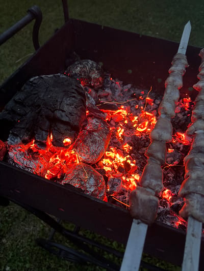 The image captures a close-up, high-angle view of a barbecue grill filled with glowing red embers, charred wood, and food items. Two metal skewers, each loaded with pieces of raw meat, are positioned horizontally over the heat source. Some of the meat appears to be marinating. In the foreground, a large piece of charred wood dominates the left side of the grill, while several foil-wrapped food items, likely potatoes, are nestled amongst the coals. The background shows a blurred green grassy area, suggesting an outdoor setting. The overall scene depicts food preparation on a barbecue grill, with the vibrant glow of the embers indicating an active cooking process. There are no people visible in the image. The location is identified as SNT Manikhino, Russia.