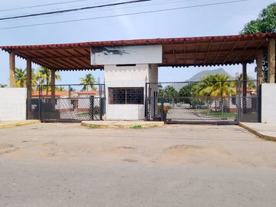 The image shows the gated entrance to a property in Aricagua, Venezuela. The entrance has a red-roofed structure supported by wooden pillars, and a central white building with a barred window. Black metal gates extend on either side of the building. A partially obscured sign is affixed to the white building, with faint lettering suggesting "El Palmar". The road in front of the gate is paved but shows signs of wear and tear. Palm trees are visible within the property behind the gate, and a distant mountain can be seen in the background. Power lines run above the structure.