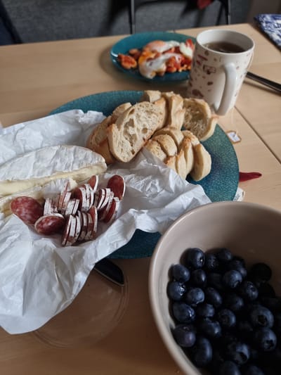The image depicts a food arrangement on a light brown surface. A white mug with a design sits near a small, blue plate containing what appears to be a seafood salad. A larger blue plate holds slices of baguette, brie cheese, and sliced sausage arranged in white parchment paper. A beige bowl filled with blueberries sits near the bread. A clear plastic container is also visible. The location in the city of Clichy, France, cannot be verified from the image. There are no people visible in the image.