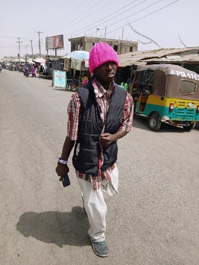 An individual is depicted walking on an asphalt road in a bustling urban street in Masvingo, Zimbabwe. The person, identified as a male, is in the foreground, moving towards the left side of the frame. He wears a bright pink knitted beanie, a red and white plaid long-sleeved shirt, a black padded vest, light-colored loose-fitting trousers, and dark grey Crocs-style shoes. A multi-strand white and black beaded bracelet is on his left wrist. He holds a dark-colored smartphone in his right hand, while his left hand is positioned near his chest, adjusting the vest. His shadow is cast on the road to his left, indicating sunlight from the upper right.

In the midground and background, the street is lined with various market stalls and temporary structures featuring corrugated iron roofs, tarpaulins, and at least one light blue umbrella. Multiple individuals are visible in the distance, engaged in activities consistent with a market environment. Utility poles with overhead power lines span the street. A large billboard with partial text "100%" is visible above the left-side structures. On the right side of the road, a three-wheeled motorized rickshaw (tuk-tuk) with a yellow, green, and brown color scheme and registration "PTA 4749" is parked. Behind the street vendors and the rickshaw, a two-story concrete block building, possibly under construction or derelict, is visible. The sky is clear and bright, consistent with a sunny day.