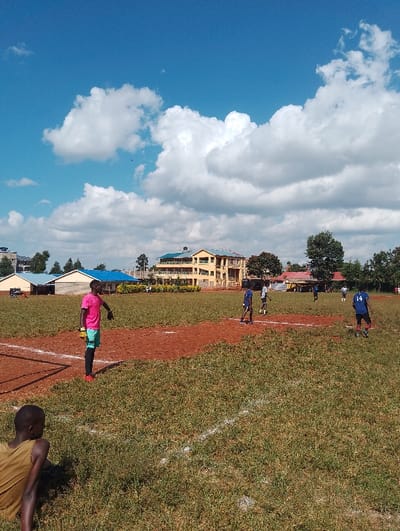 The image depicts a group of young men playing soccer on a field in Rungiri, Kenya, at the coordinates -1.249 latitude and 36.691 longitude. The field is covered with short green grass with white lines painted across it. There are also areas of red dirt visible. The players are wearing soccer uniforms, and one is wearing a pink shirt and teal shorts. The image is taken from a slightly elevated angle looking down on the field. The men are facing away from the camera, and one is looking towards the camera. Behind the players, there is a building with a yellow facade and a blue roof. The building is in the middle distance and has a few trees growing around it. The sky is clear blue with white clouds, and the sun is shining.  The background of the image includes a few scattered trees, a few buildings, and a blue sky.  The image is taken in the daytime. There are a few people on the field, including the soccer players, and a man sitting in the grass in the foreground. There is a person in the background, to the right of the soccer players.
