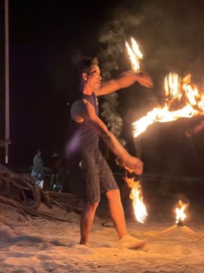 An adult male fire performer is depicted mid-action on a sandy beach at night in Kamala, Thailand. The performer, wearing a dark tank top and dark shorts, is barefoot and positioned with his body in motion, suggesting a spin or sweep. He holds and manipulates at least two fire props, one in each hand, which produce bright orange and yellow flames, emitting visible smoke. His face, illuminated by the fire, is seen in profile, with a focused expression. To the immediate right, a smaller, stationary fire torch burns on a low sand mound. In the foreground on the left, exposed, gnarled tree roots or driftwood are partially buried in the sand. Further left, a vertical wooden post is visible. The background is dark, indicating nighttime, with blurred figures of other individuals observing the performance. The primary illumination for the scene originates from the burning fire props and torches.