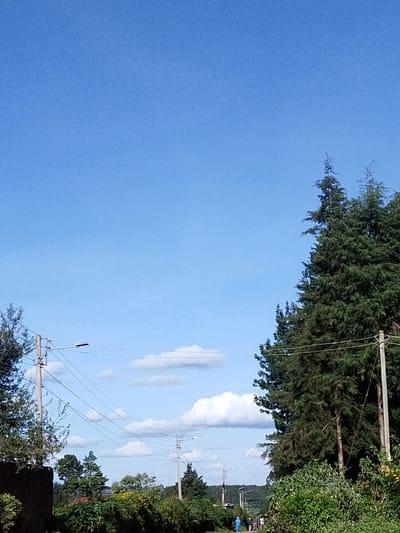 The image depicts a landscape in Bibirioni ward, Kenya. The sky is predominantly blue with scattered white clouds. Electrical poles with connecting wires run across the scene. Vegetation, including trees and shrubs, fills the lower portion of the image. A group of people is visible in the distance along a pathway. The environment appears to be rural or suburban.