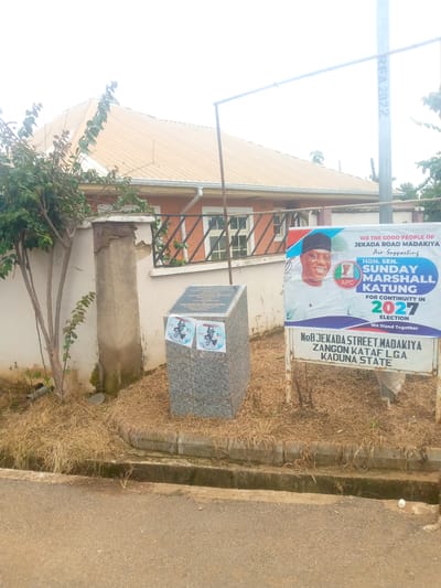 The image captures a street scene in Madakiya, Nigeria. Dominating the right side of the frame is a large campaign banner for "Hon. Sen. Sunday Marshall Katung" of the APC party, advocating for his re-election in 2027. The banner displays a smiling portrait of Katung and the text "WE THE GOOD PEOPLE OF JEKADA ROAD MADAKIYA Are Supporting" and "FOR CONTINUITY IN 2027 ELECTION We Stand Together." Below this banner is a sign indicating the address: "N08 JEKADA STREET, MADAKIYA, ZANGON KATAF LGA, KADUNA STATE."
To the left of the banner, a grey granite monument stands, adorned with two smaller circular posters featuring images of individuals, likely political figures, and the APC logo. The monument appears to be a memorial or information plaque, though the text on it is difficult to read clearly. A bush with green leaves grows to the left of the monument and the wall it is situated against.
The environment consists of a paved road in the foreground, a dry, overgrown grassy area surrounding the monument and banner, and a building with a corrugated brown roof in the background. A concrete wall with peeling paint and a black metal railing borders the property where the monument and banner are placed. A utility pole with some markings, possibly "REA 2022," is visible on the far right. The sky is overcast.