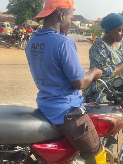 This image captures a scene in Galadimawa Village, Nigeria. Two individuals are prominently featured, seated on a motorcycle. The person in the foreground, wearing an orange cap and a blue shirt, is looking to the side. Their shirt displays text that reads "APO SHOPPRENDERS" followed by a series of numbers: "01146379", "355167031". The second person, seated behind, is wearing a blue headscarf and a blue and yellow patterned traditional garment. The motorcycle is red and black.
In the background, a row of other motorcycles is visible, suggesting a gathering or transport hub. A yellow kiosk-like structure and some umbrellas are also present, indicating a market or roadside vendor area. The overall impression is that of a typical day in a Nigerian village, likely during daylight hours, with a warm, possibly sunny, atmosphere. The image appears to be taken from a slightly elevated angle, looking down at the subjects and the surrounding scene. The focus is primarily on the two individuals and the motorcycle they are on, with the background providing context to their location and the potential activities occurring around them.