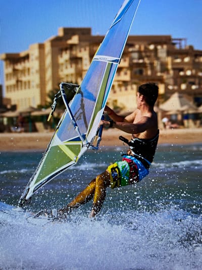 A windsurfer is captured mid-maneuver in Bacong, Philippines, with buildings visible along the coastline behind. The windsurfer, a man wearing a black tank top and colorful shorts, is leaning forward on a windsurf board with a triangular sail that is blue, green, and white. The board is cutting through the water, creating a splash of white spray. The sky is clear blue, and the buildings are beige in color.
