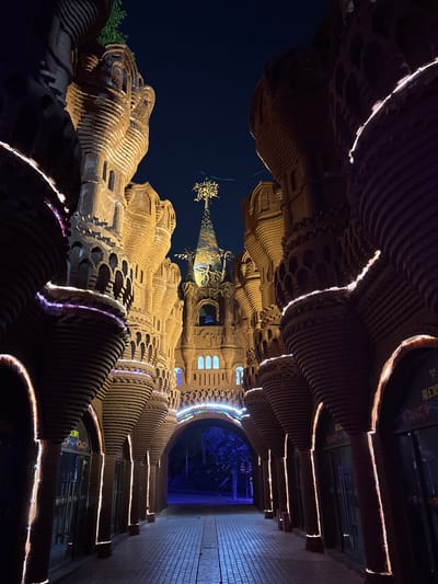 A low-angle shot captured at night in Baqiao, China, showcases a whimsical architectural structure that bears a resemblance to Antoni Gaudí's Sagrada Familia. The structure, built from stacked, undulating layers of material, is illuminated with warm, ambient lighting that accentuates its contours and details. The camera angle gives a perspective of an archway leading to a blue-lit tunnel. The sky above is dark. The structure appears to be situated within an entertainment or commercial area, given the signage visible along the archway.