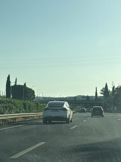 The image shows a view from a vehicle driving on a highway, likely in or near Oeiras, Portugal, given the context of other images from this set. The sky is clear and pale blue, with faint horizontal lines possibly indicating power lines or the horizon. On the right side of the highway, there are green bushes and trees, with some tall, slender trees that appear to be cypress trees. Beyond the bushes, there is a glimpse of buildings and possibly a bridge structure.
In the foreground, the asphalt highway is visible with white lane markings. A silver Tesla Model X is driving in the right lane, with its rear clearly visible. The car has a license plate with Greek characters, suggesting it may be a foreign-registered vehicle in Portugal, or the image was taken elsewhere. Further ahead in the same lane, a dark-colored SUV is visible. In the left lane, a black SUV is also visible. The overall scene depicts a typical highway driving scenario with several vehicles on a clear day.