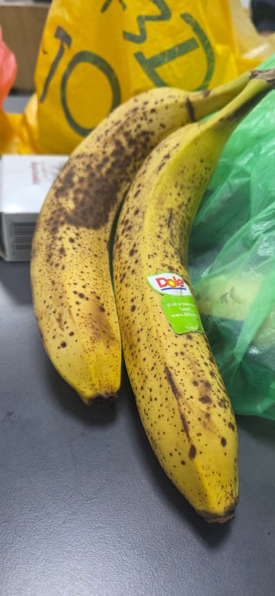 The image shows two ripe bananas with brown spots on a gray surface. They appear to be resting on or next to a partially visible green plastic bag and a yellow grocery bag. One of the bananas has a Dole sticker on it. The image does not contain any information related to the city of Drinovo, Bulgaria.