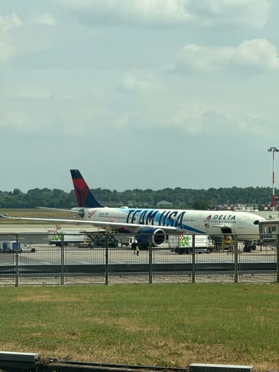 An outdoor, daytime image captures a Delta Airlines aircraft parked at an airport gate in Ferno, Italy. The prominent subject is a white Airbus A220-300 featuring a distinctive "TEAM USA" livery in large blue text across its fuselage, with the standard "DELTA" logo also visible near the cockpit. The aircraft's tail displays the iconic red, white, and blue Delta Airlines logo, and its registration number, "N471DX," is clearly visible.
The airplane is situated on a paved tarmac, with a jet bridge partially visible connecting to its front. Various ground support vehicles, including catering trucks (one showing "gourmet" text), baggage carts, and other service equipment, are positioned around the aircraft, suggesting active ground servicing or preparation for a flight. A metal security fence runs along the edge of the tarmac, separating it from a section of green grass in the foreground.
In the background, a lush line of green trees forms a natural barrier. The sky above is overcast, with soft, diffused clouds, indicating a cloudy day. The scene depicts a typical airport operation, with the special livery of the Delta aircraft being a notable detail.