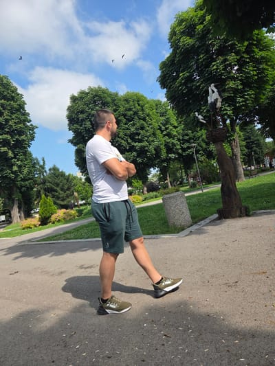 The image depicts a man standing in a public park in Elin Pelin, Bulgaria, on a clear, sunny day. The man, with short dark hair and a beard, is light-skinned and wears a white short-sleeved t-shirt, dark green athletic shorts with a drawstring, and olive green athletic shoes with "PUMA" branding. He stands with his arms crossed, facing right and looking slightly upward. He is positioned on a dark gray paved pathway, which shows scattered small debris. To his right, on a patch of green grass, is a cylindrical concrete trash receptacle. Further right, a prominent wooden sculpture featuring two adult white storks and one younger stork in a nest, mounted on a carved wooden tree trunk, stands beneath a large deciduous tree. Numerous other large deciduous trees with dense green foliage populate the park, casting shadows on the ground. In the background, to the right, other park-goers are visible, along with park benches. To the left of the paved path, manicured bushes with pink and yellow flowers are visible, along with more trees. The sky above is blue with scattered white cumulus clouds, and several small dark birds are visible flying.
