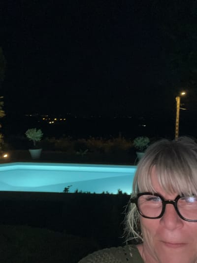 The image shows a nighttime scene with a woman in the foreground wearing glasses. Behind her is a bright blue swimming pool, and beyond that, a dark landscape with distant lights that suggest a town or city. There are also trees and a lamppost visible. The overall impression is that of a residential area in or near Saint-Galmier, France, at night, with the swimming pool illuminated.