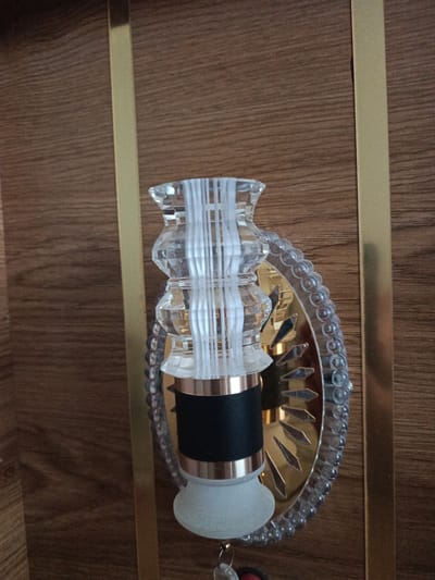 The image shows a close-up of a decorative element, possibly a light fixture or part of a piece of furniture, against a wooden background. The object features clear, textured glass with vertical ribbed detailing, a black cylindrical section, and a gold, fan-shaped reflective plate with diamond-shaped cutouts. A beaded clear plastic trim surrounds the gold plate. A metallic gold vertical strip runs along the right side of the wooden surface. The overall setting appears to be indoors, with the wood grain suggesting a finished surface such as a door or paneling. There are no people or discernible activity in the image. The location within Hakkulabad, Uzbekistan, is not evident from the visual information provided.