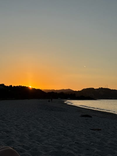 The image depicts a serene beach scene at sunset in the Municipal Unit of Kassandra, Greece. The sun, partially obscured by a silhouetted treeline on a hill, casts a warm, orange glow across the sky, transitioning to a pale blue towards the top. The calm sea reflects the sunset colors, with gentle waves lapping the shore. The foreground is dominated by dark, textured sand, showing ripples from the wind or water. In the mid-ground, several indistinct figures are visible on the beach, some appearing to be walking or standing. A few cars are parked near the treeline on the left side of the image. The overall impression is one of tranquility and natural beauty.