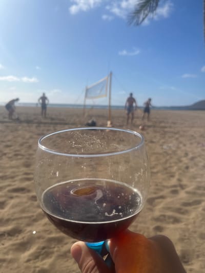 The image, taken at Las Canteras Beach in Las Palmas De Gran Canaria, Spain, shows a hand holding a wine glass filled with red liquid, obscuring part of the view. In the background, several people are playing volleyball on the sandy beach. A volleyball net is visible in the mid-ground. The sky is partly cloudy.