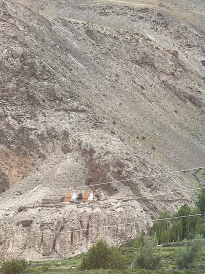 The image shows a mountainous landscape with a rocky slope and scattered vegetation. In the mid-ground, several stupas, traditional Buddhist shrines, are visible on a rocky outcrop. These stupas are painted in varying colors, including white, orange, and black. A stone structure is also present near the stupas. The foreground consists of a grassy area with trees and bushes. Several power lines traverse the image horizontally. The environment appears to be dry and arid, characteristic of a mountainous region. The image is taken in the vicinity of Char, India, a location known for its monasteries and ancient structures.
