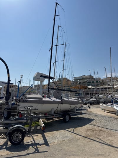 The image shows a marina in Anzio, Italy on a clear day. In the foreground, a sailboat is mounted on a trailer, covered in a grey tarp. The sailboat has a tall, black mast extending upwards. The trailer is parked on a paved surface with cobblestone patterns. The shadows of the mast and rigging are cast on the ground. In the background, there are several other sailboats berthed at the marina. Buildings are visible behind the masts of the other boats. The sky is clear and blue. No people are visible in the shot.