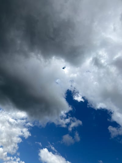 The image shows a sky with varied cloud formations and blue sky visible. In the upper half of the frame, dark grey storm clouds dominate, with some lighter grey and white clouds interspersed. Towards the center of the image, a small, dark object, possibly a drone or a bird, is visible against a patch of lighter sky. The lower half of the image transitions to a brighter blue sky with scattered white cumulus clouds. The image was taken in Jodłownik, Poland.