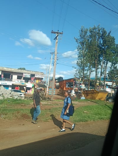 The scene is a street in Karingani ward, Kenya, under a partly cloudy sky. A wooden utility pole with power lines is in the center. A man wearing a dark shirt, blue jeans, a gray cap, and hiking boots walks on a dirt road. A young woman in a light blue school uniform, white socks, black Mary Jane shoes, and a dark blue bag crosses the road. Buildings with storefronts are on the left and in the background, with signs reading "SIR-G STUDIOS", "MOH SALON", and "INDAGANI TRADERS". A white car is parked near a pile of stones. A yellow dumpster is on the right side of the road, near a patch of grass, with a partially visible black car. A shadow stretches from the lower right corner into the scene.