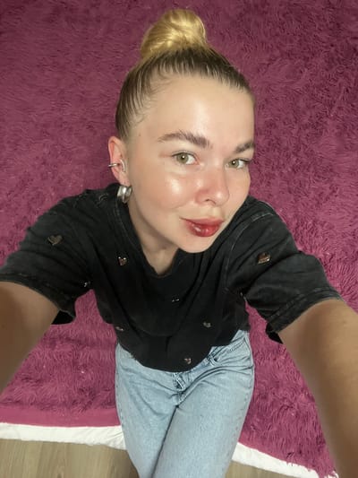 A young woman with blonde hair in a bun, wearing a black t-shirt with heart accents and light blue jeans, is captured from a high angle in a selfie. She has green eyes, full lips with red lipstick, and is wearing hoop earrings. The woman is positioned on a soft, pink, furry surface, likely a rug or blanket, which is spread over a white bedspread. The lighting suggests an indoor setting. This image does not contain any discernible information that would allow for its localization to the city of Tikhoretsk, Russia.
