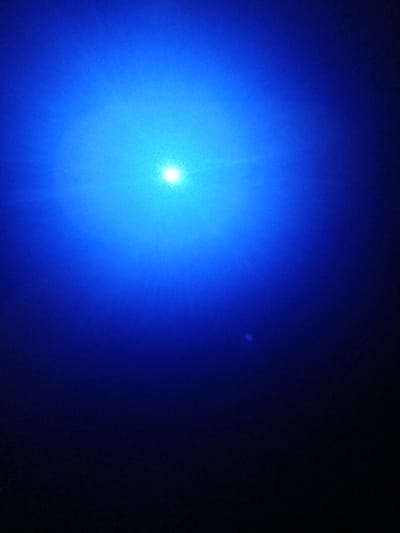 A bright, circular, and intensely blue light dominates the left side of the image, radiating outwards with diminishing intensity. Its center is a sharp, white point. The background is a gradient of dark blue to almost black, transitioning towards the right edge. A small, faint, circular spot is visible in the upper right quadrant, contrasting with the darkness surrounding it. The overall scene evokes a sense of deep space or a powerful light source against a dark backdrop.