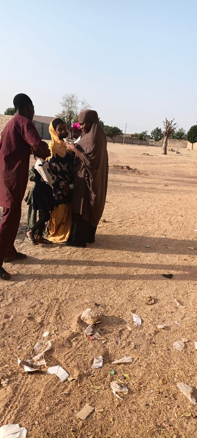 Four people stand in a sandy area with litter scattered on the ground. One man in a maroon shirt stands to the left, while a woman in a brown hijab stands to the right. Between them, a young girl in a black and yellow patterned dress and a yellow headscarf looks on. The other person is another young girl wearing a pink headscarf and standing behind the girl in yellow. A few trees and buildings are visible in the background under a clear sky.