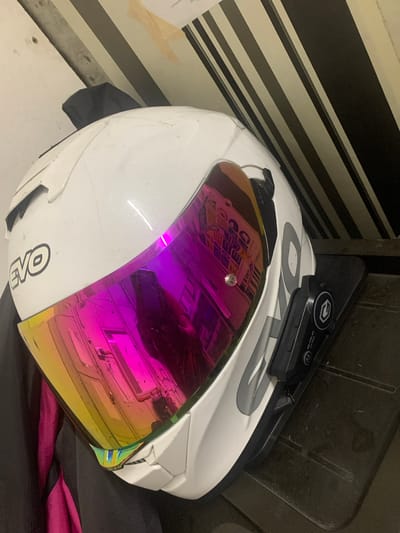 An angled, close-up, eye-level, indoor shot shows a white motorcycle helmet with a reflective, iridescent visor. The helmet has a prominent "EVO" logo on the side and a dark-colored attachment, likely a communication device, mounted on the right side. The helmet rests on a dark, textured surface, possibly a table or shelf. Behind the helmet, a wall displays a striped pattern of black and off-white bands. There is a piece of yellow tape visible on the wall above the helmet. The scene appears to be in a residential setting in Angeles City, Philippines.