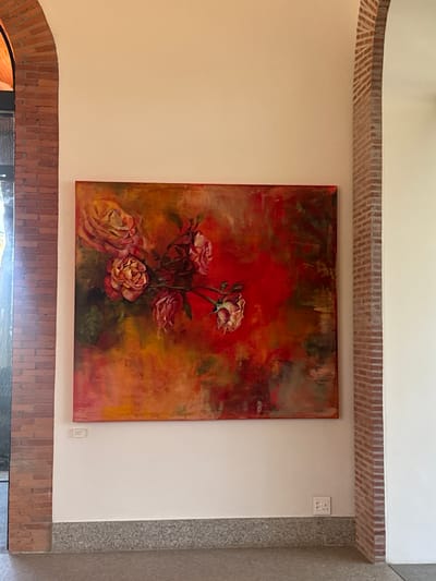 A square painting of a floral arrangement hangs on a cream-colored wall, situated between two brick lintels. The painting features a mix of red, orange, and green hues, with flowers depicted in shades of pink, peach, and cream. An electrical outlet is visible above the painting on the right. The wall is flanked by a textured grey column on the right, suggesting an interior space. The scene's location is identified as Stellenbosch Nu, South Africa.