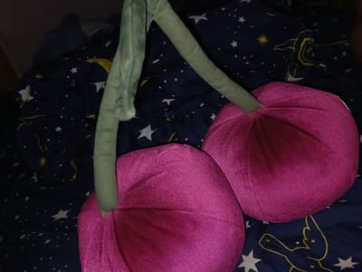 The image shows two plush cherries with green stems lying on a dark blue fabric with a pattern of stars and moons. The cherries are a vibrant pink-purple color and appear to be made of a soft, velvety material. One cherry is larger and positioned in the foreground on the left, while the other is slightly smaller and to its right. The stems are thick and fuzzy green. The background fabric is dark blue with white stars of various sizes and some yellow celestial elements, possibly a moon or a stylized sun. The lighting is dim, suggesting it may be nighttime or an indoor setting with low light. There is no visible indication of the location being in El Espinal, Venezuela, or any specific city. The scene is static and focuses solely on the plush toys.
