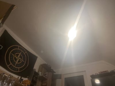 The image depicts an indoor scene, likely a bedroom or living area. A bright light fixture, possibly a ceiling light, is prominently visible, emitting a strong glare that washes out parts of the upper portion of the image. On the left side, a black flag with a circular logo, appearing to be a Stone Island compass badge, hangs on the wall. Below the flag, a shelf or surface holds several plastic water bottles and other indistinct objects. To the right of the flag, a portion of a wooden shelving unit or cabinet is visible, with more objects on its shelves. Further to the right, a doorway or opening leading to another room is partially visible, and above it, the top of a piece of furniture or closet is seen. The overall lighting suggests it is either nighttime or the room is otherwise dimly lit, with the ceiling light being the primary illumination source. No individuals are present in the frame. The image does not contain any specific visual cues that would allow for identification of the location within Gelsenkirchen, Germany.