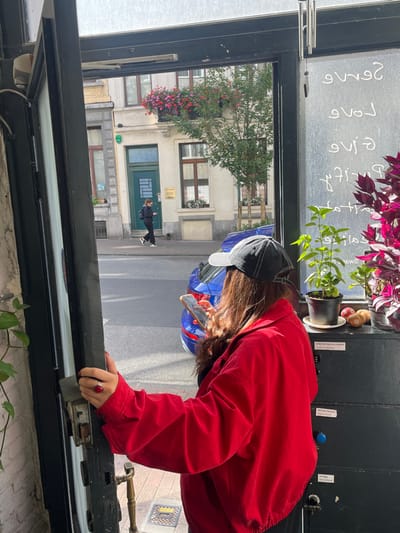The image depicts a scene observed through an open doorway from inside a building on Rue de Mérode 27, 1060 Saint-Gilles - Sint-Gillis, Brussels, Belgium.
In the foreground, a female subject is viewed from the rear, positioned within an open, dark-framed doorway. She has long, dark brown hair, wears a red long-sleeved jacket, a dark garment, and a dark baseball cap with a light-colored brim. A large red ring adorns her left ring finger. She holds a light-colored smartphone with both hands, viewing its screen. Her right hand rests on the interior door frame near a metal lock mechanism.
To the subject's right, within the interior, a black multi-drawer cabinet is visible. On its top surface are two potted plants—one with green leaves, another with magenta foliage—and several pieces of fruit. A label on the cabinet reads "UNEUM TRANSLATIONS." A glass panel to the right of the doorway displays backward white text, including "Serve Love Give," which are reflections from inside the establishment. A metallic wind chime hangs from the top of the door frame. A green potted plant is partially visible on the left interior side.
Beyond the doorway, an asphalt street with a parked blue car (rear section visible) extends into the mid-ground. Across the street, a second female subject walks rightward on the sidewalk, wearing dark clothing and white-soled sneakers, and also viewing a smartphone. The background features light-colored, multi-story European buildings. The most prominent building directly opposite has a distinctive green entrance door with two small golden plaques mounted above it. Its first-floor balcony is adorned with vibrant red and pink flowers in window boxes. A small deciduous tree with green leaves is planted on the sidewalk, supported by wooden stakes. The scene is illuminated by bright, diffused daylight.