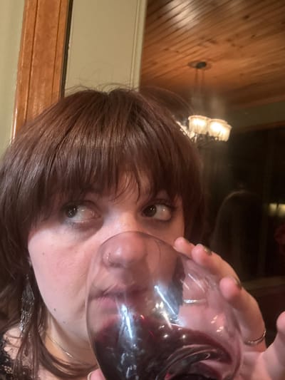 A woman holds a glass of red wine up to her face, nearly touching her nose. She has brown hair with bangs. A wood paneled ceiling is visible in the background, along with a chandelier-style light fixture. The woman wears a silver necklace. Her fingernails are dark. She is looking directly at the camera.