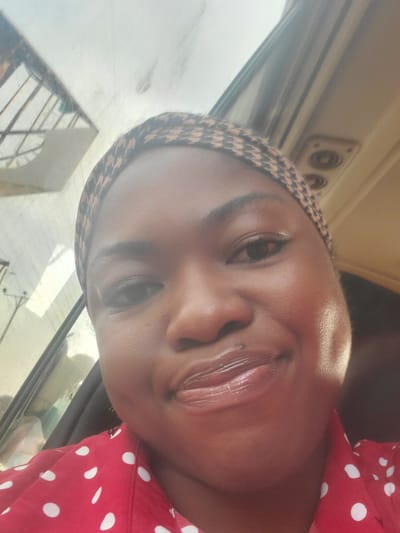 A young Black woman is taking a selfie inside a vehicle. She is wearing a red shirt with white polka dots and a patterned headscarf. Her head is slightly tilted, and she is smiling. The interior of the vehicle is visible, including a window with a reflection and the ceiling with a vent. The background suggests an urban environment, potentially Kakau, Nigeria, given the context.