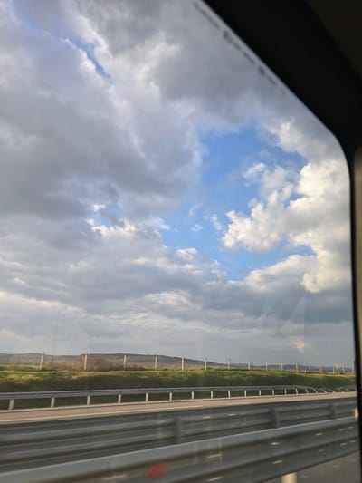 The image, captured from inside a vehicle, presents an exterior view of a highway landscape near Yazdach, Bulgaria. The upper two-thirds of the frame show a dynamic sky with a mix of light and dark grey clouds, interspersed with clear blue sky and bright white cumulus clouds, particularly in the upper right and center. Below the sky, a distant, undulating horizon line reveals green and brown fields or hills. In the midground, a green grassy verge runs parallel to the road, featuring a line of regularly spaced, light-colored fence posts with wires strung between them. The foreground displays a multi-lane asphalt highway, characterized by two parallel sets of grey metal guardrails; the lower set is closer to the viewer, while the upper set runs along the edge of the grassy verge. Faint white lane markings are visible on the road surface. No people or other vehicles are present in the observable scene.