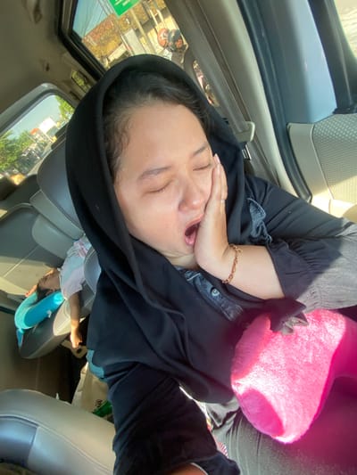The image depicts a woman in a black hijab yawning inside a vehicle, her hand pressed against her cheek. In the background, a child sleeps on a blue pillow in the car's back seat. The car appears to be in motion, with a view of a street scene through the windows, suggesting a location in the city of Bangsal, Indonesia, possibly on a sunny day. Buildings, trees, and other vehicles are visible outside. A bright pink cushion is held in the woman's lap.