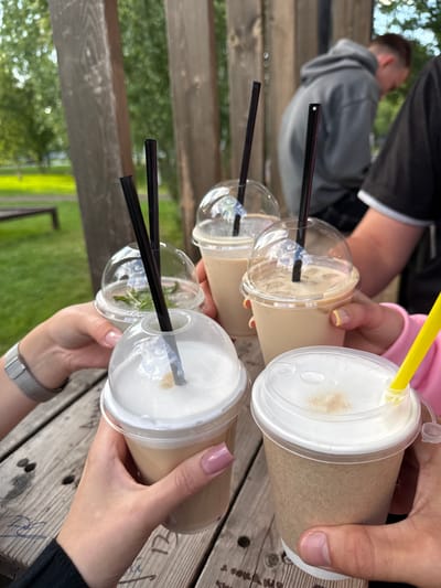 Four people are holding iced coffee drinks in clear plastic cups with black straws. The drinks are on a wooden table. In the background, two men are visible, one wearing a gray hoodie and the other a black t-shirt. The environment appears to be outdoors, with trees and greenery visible. The location is identified as a cafe in Mozhga, Russia. The image captures a moment of social gathering and enjoyment of beverages.