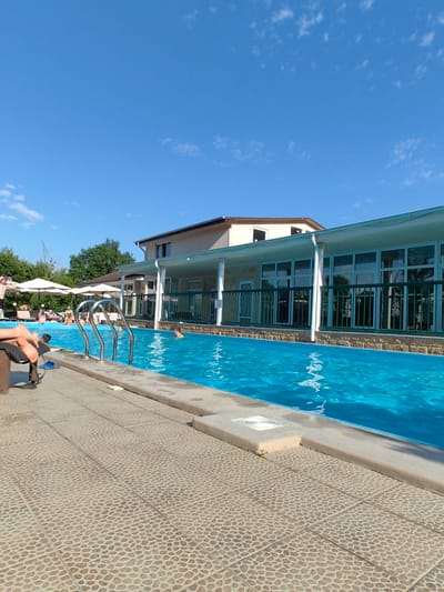 A bright, sunny day at an outdoor swimming pool. The pool is filled with clear blue water and has a set of metal stairs leading into it. The poolside is paved with a tan pebble-like tile. In the background, a two-story building with a light-colored facade and a green railing is visible. There are several people in and around the pool, some swimming, some relaxing on lounge chairs under large white umbrellas. Trees line the edge of the property. The sky is a clear, vibrant blue with a few scattered white clouds. The image appears to be taken in Oryol-Izumrud, Russia.