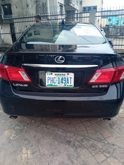 The rear view of a black Lexus ES 350 sedan is captured in this image, parked on a cobblestone surface. The car's license plate, which reads "PHC-149AT RIVERS", is clearly visible, along with the "LEXUS" and "ES 350" badging. The car has red taillights and a chrome strip above the license plate. A paper with text is adhered to the rear windshield. In the background, a rusty metal fence separates the car from a building with a weathered facade. A person, partially obscured, stands behind the fence, looking towards the car. The overall scene suggests an urban environment, possibly a street or driveway in Rumuepirikom, Nigeria.